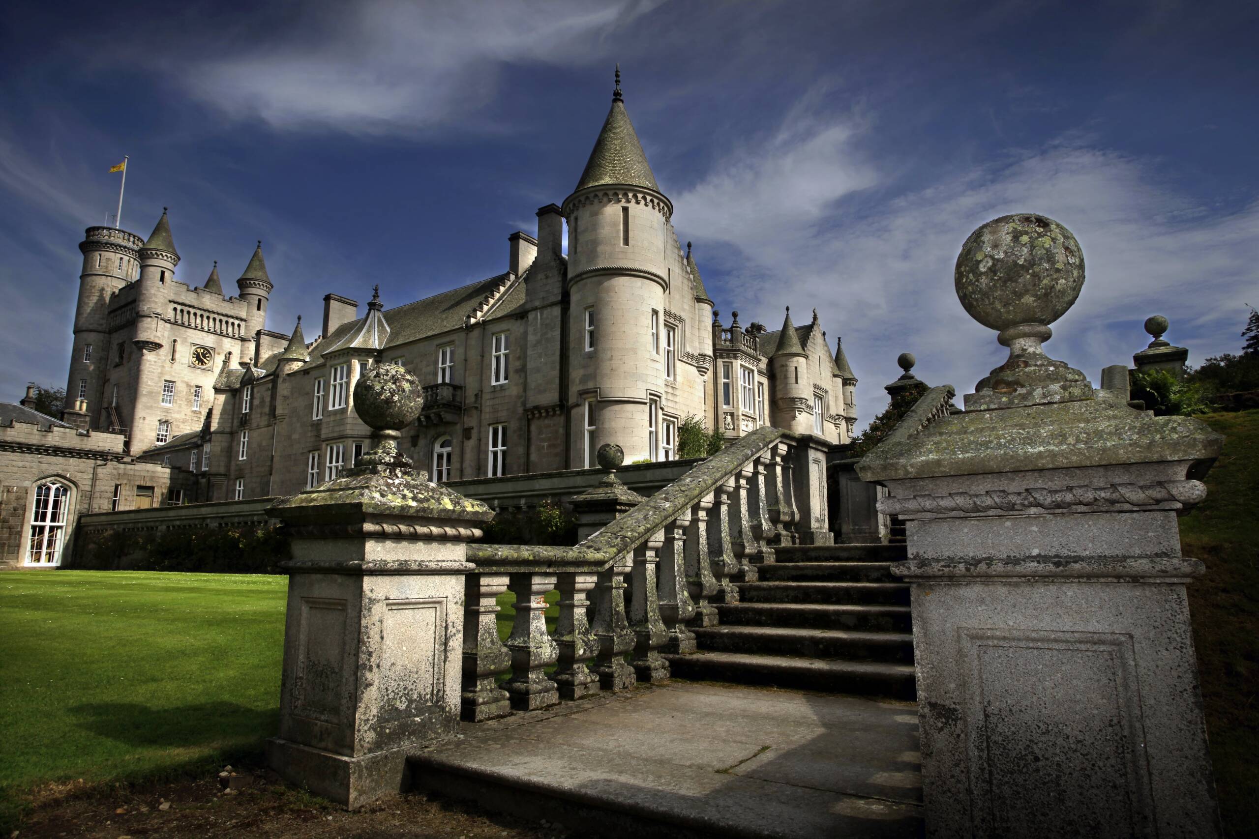 Balmoral Castle is shown up close, featuring its grand stone staircase and ornate turrets under a partly cloudy sky, with well-manicured green lawns in the foreground.